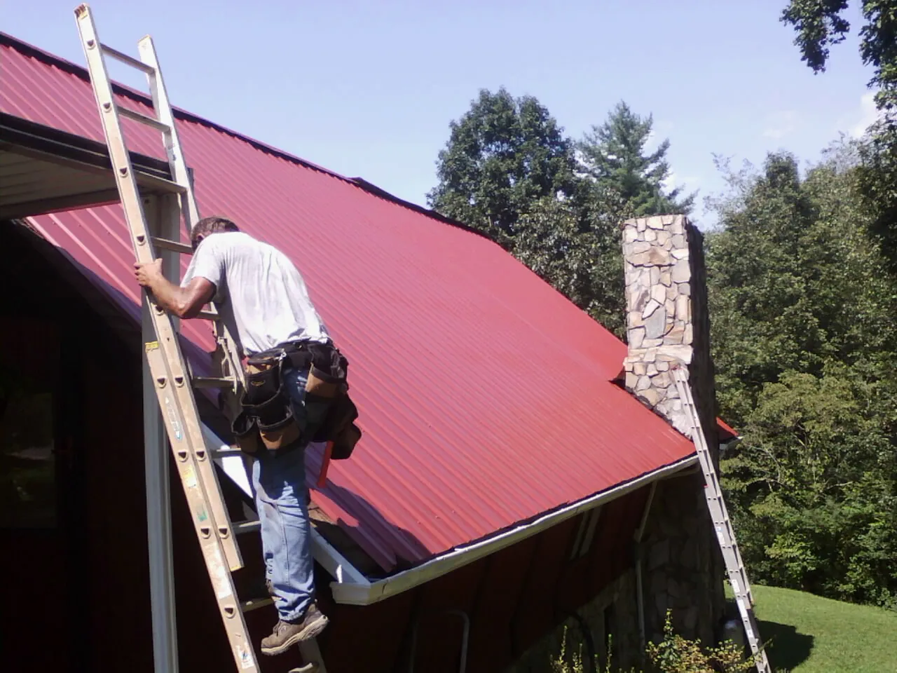 Roofer on a ladder beside a red metal roof and stone chimney — Soffit & Fascia Repair by Roofing Brothers in Elk Grove